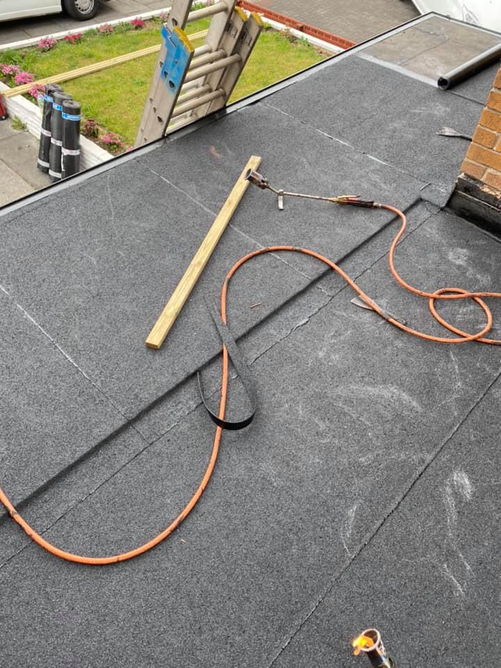 A flat grey felt roof being installed with a gas blowtorch, wooden batten, and hose lying across the surface.