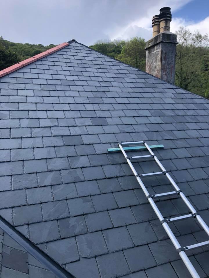 A grey slate pitched roof with a red ridge cap, an old brick chimney, and a roof ladder in place.