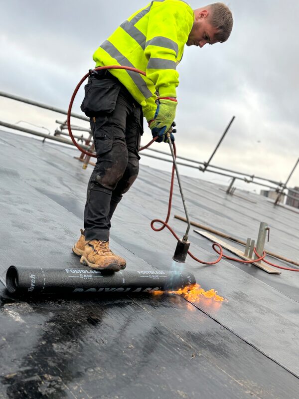 A roofer in a high-visibility jacket using a blowtorch to heat-weld a black felt roofing roll into place.